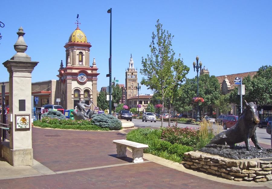 A plaza corner with a stone gate post, a bronze boar statue, flower beds and a Spanish-style tower behind