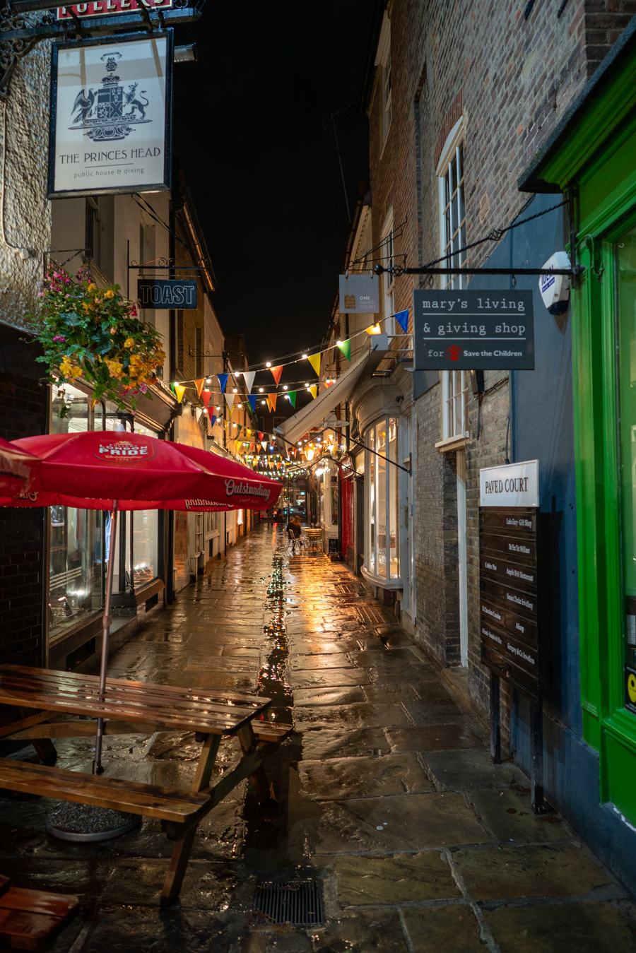 A narrow wet flagstone lane strung with bunting and lights, the Prince's Head sign on the left and a Paved Court name board on the right