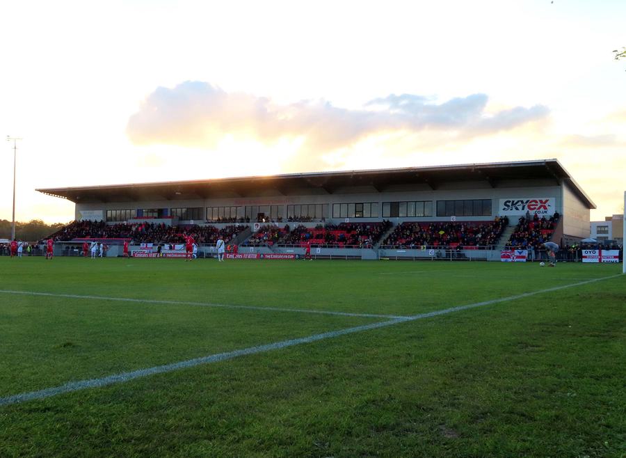 A small football ground at dusk: a single covered stand with the SKYex name along its roof, seen across the pitch