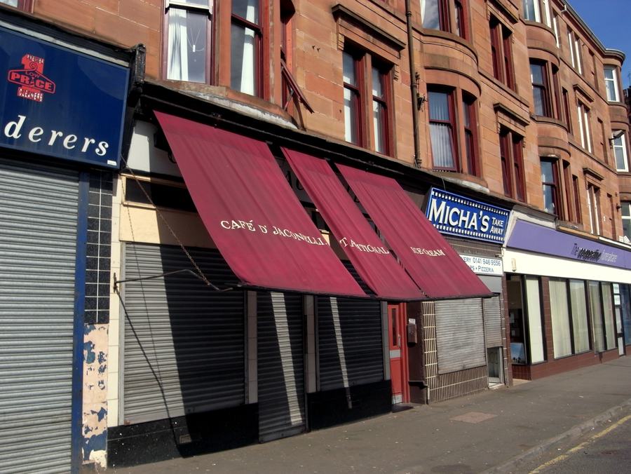 A café front with a red awning reading Café D'Jaconelli, in a red sandstone tenement, shutters half down.