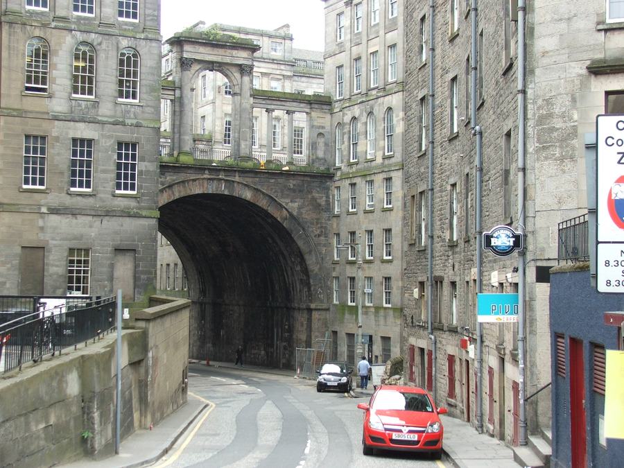 A road dipping downhill under a large stone arch between tenement buildings, cars parked along the kerb.