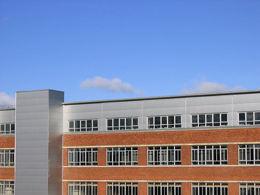 Part of a long red brick factory block with strip windows and a modern grey stair tower, under a blue sky.