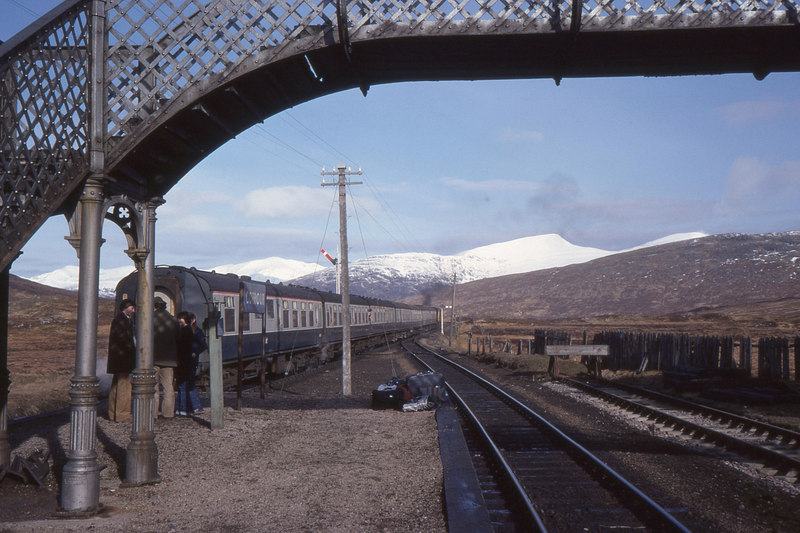 A train at a remote platform framed by the ironwork of a footbridge, snow on the hills beyond.