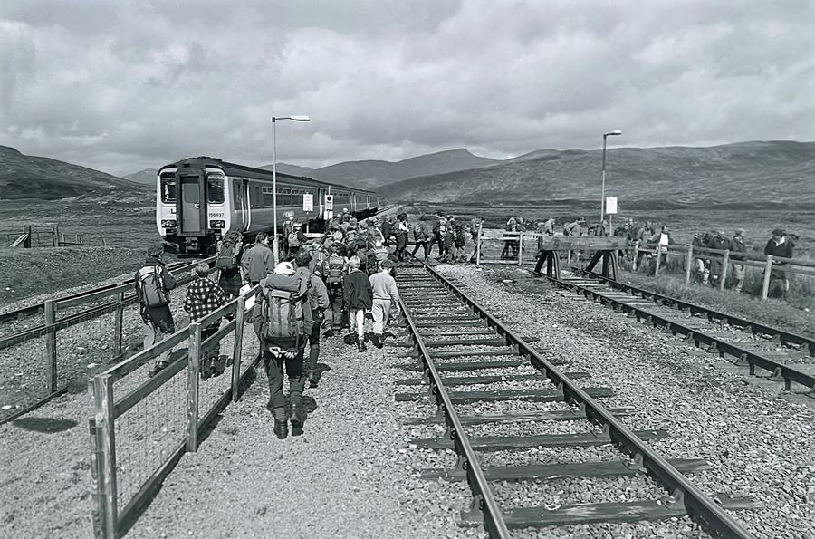 Black and white photograph of a crowd with rucksacks stepping down from a train onto a bare moorland platform.