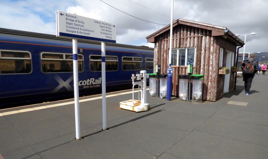 A blue and white train at a bare platform beside a small wooden shelter, walkers waiting.