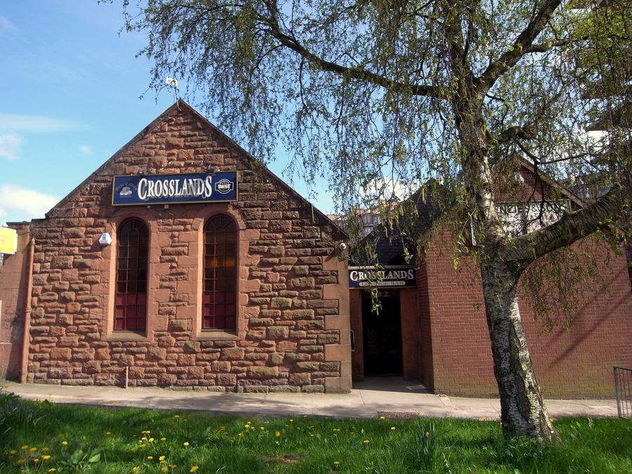 A small stone building with a gabled front and arched windows, a sign reading Crosslands above the door, trees around it.
