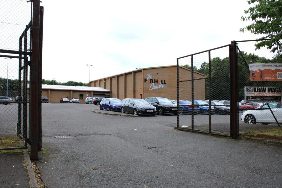 A low sports building behind steel mesh fencing and a car park, with floodlight poles.