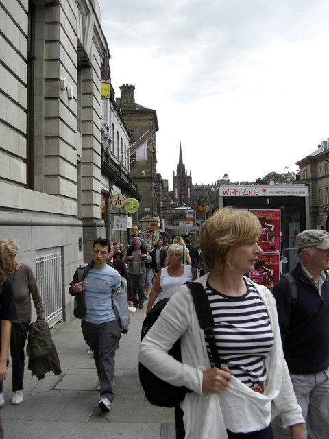 A narrow city street sloping downhill between tall stone buildings, crowded with pedestrians.