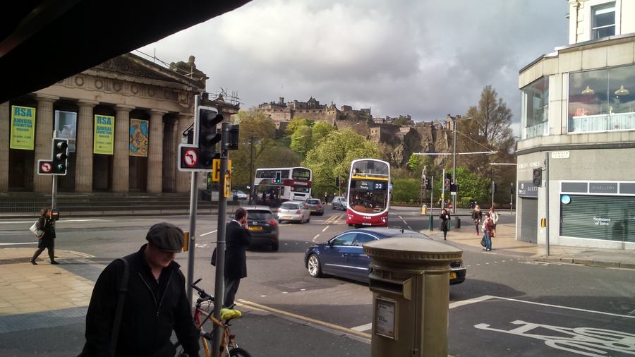 View from under a stone colonnade across a busy junction; a castle on a crag fills the skyline behind buses and traffic lights.