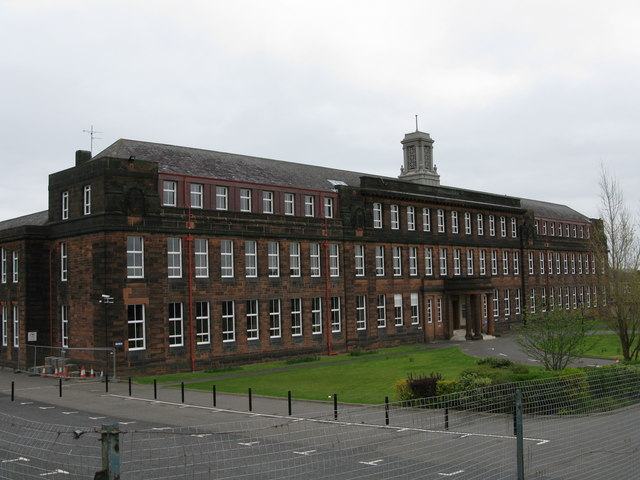 A long two-storey stone school building with a small cupola, seen across an empty playground.