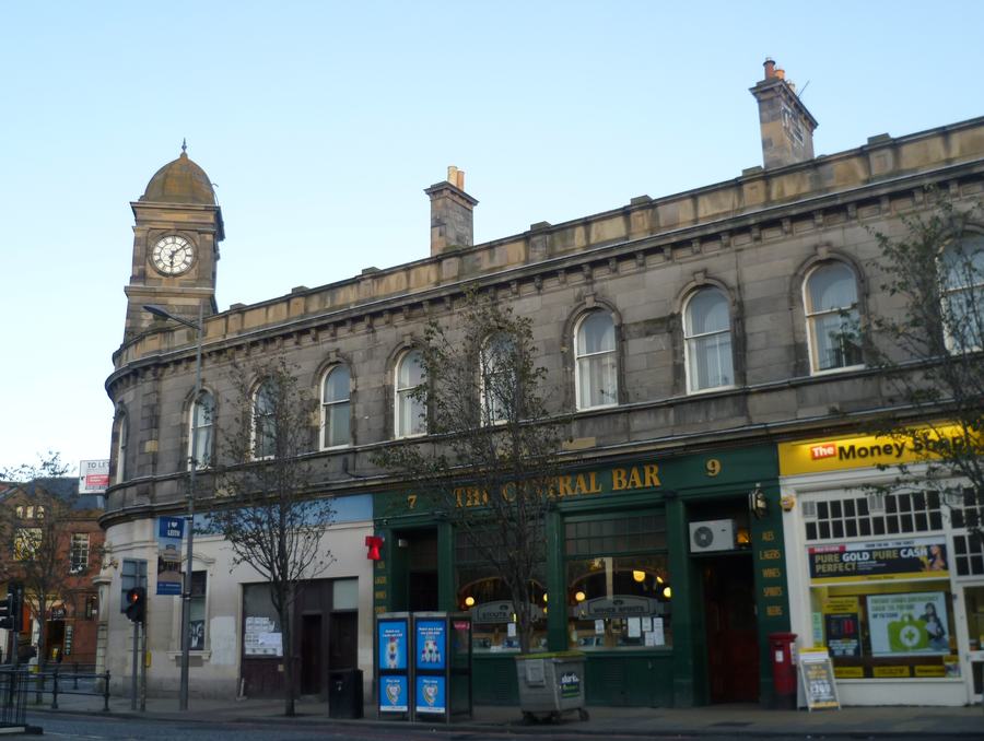 A long two-storey stone building with round-headed windows and a clock tower, a green-fronted pub at street level.