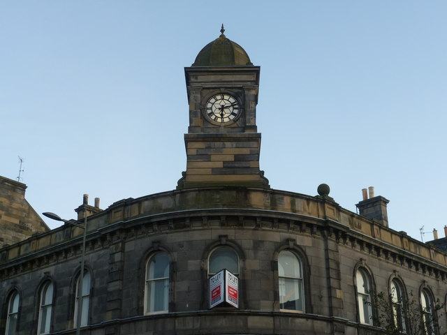 A square stone clock tower with a domed cap rising above a corner building against a blue sky.
