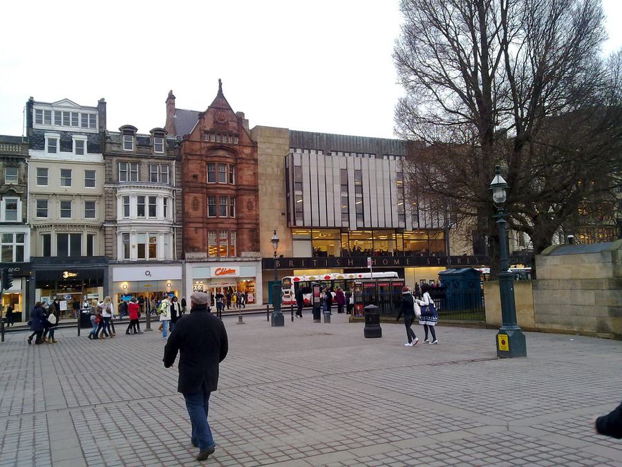 Wide paved street with chain-store fronts, shoppers walking, a red sandstone building on the left.