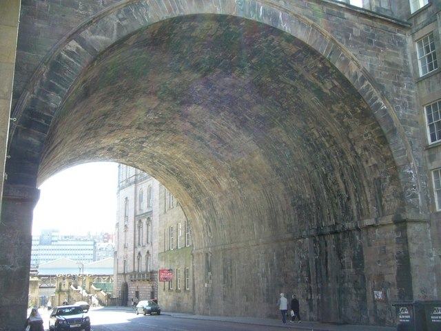 Looking up into a wide stone archway carrying a road overhead, daylight and buildings beyond.