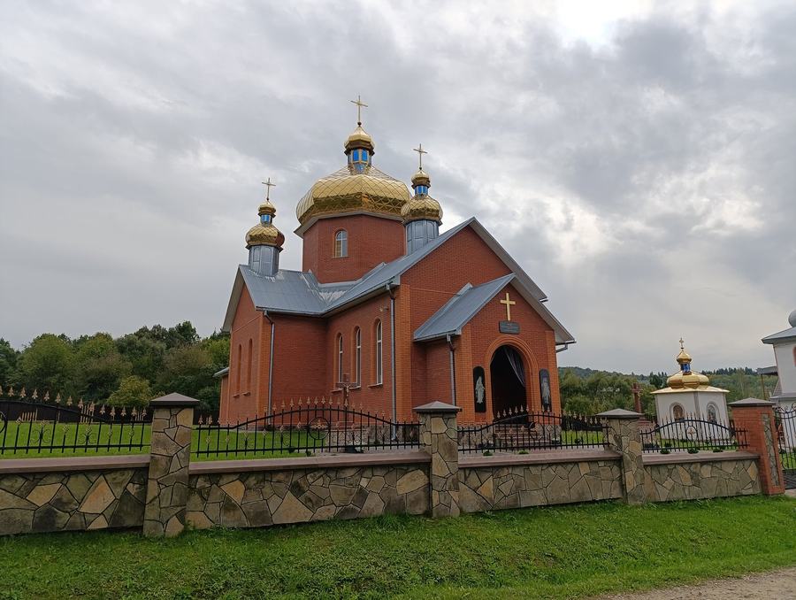 A red brick church with gilded domes behind a stone and iron fence under a grey sky