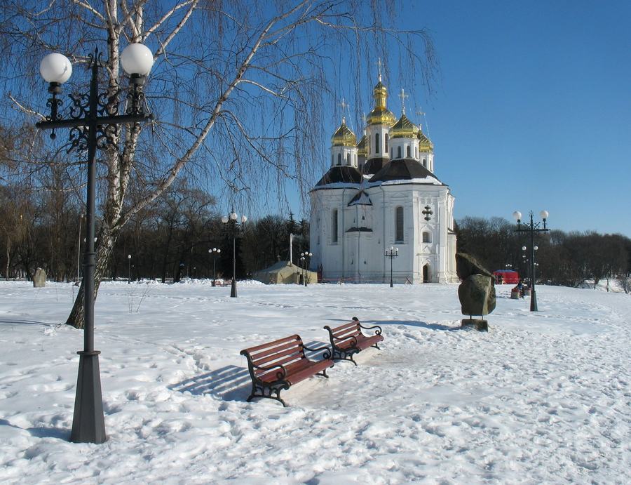 A white baroque church with five gilded domes across a snow-covered park with benches