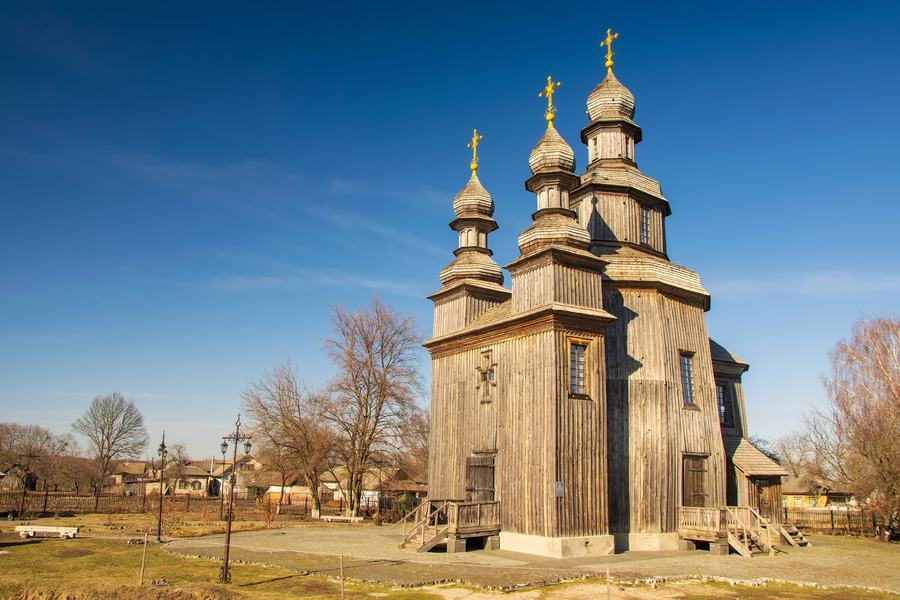 A three-domed wooden church of dark grey timber against a blue sky, low houses behind it