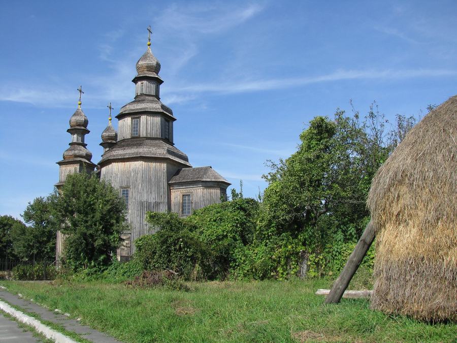 A dark wooden church with three onion domes, a large round haystack in the foreground