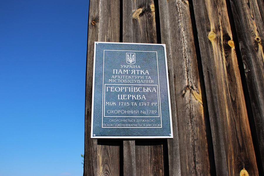 A grey metal heritage plaque with Ukrainian lettering screwed to weathered vertical planking