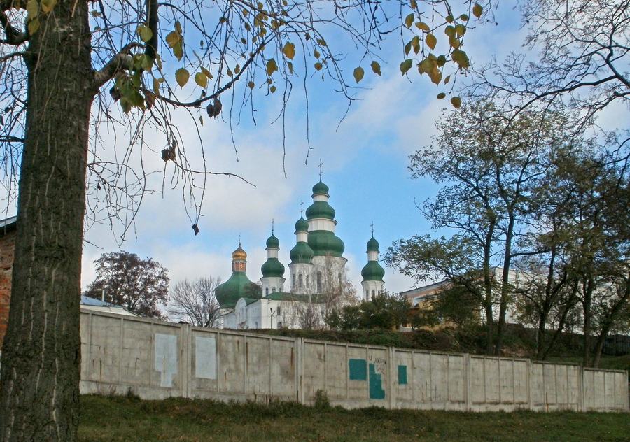 Green and gold domes of a white masonry cathedral rising above a concrete fence and autumn trees